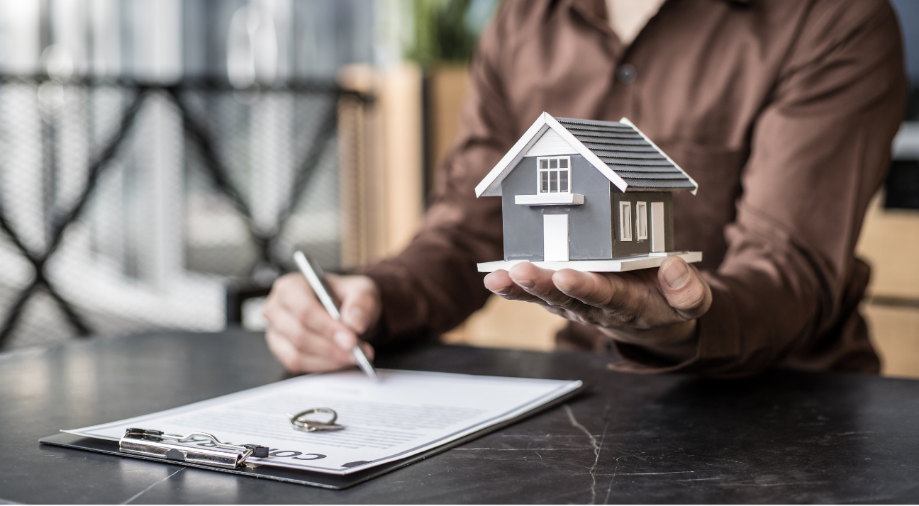 Agent holding a model house while a client signs a contract